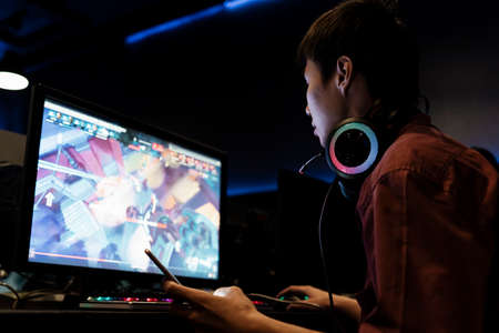 Asian gamer playing computer game at internet cafe, wearing headphone, looking at monitor screen.