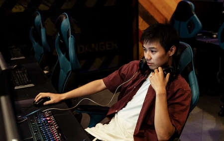 Young professional asian gamer sitting on a chair, removing headset on his head, playing PC game at internet cafe.の写真素材