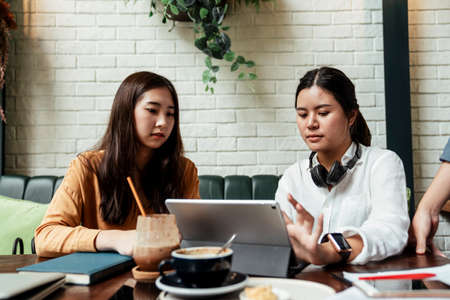 Two young ladies working together on tablet while her boss is watching. Modern technologies in daily life. Two businesswomen employees colleagues discussing working together on laptop, explaning new online idea for their startup business.の写真素材
