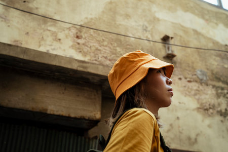 Low angle shot of yellow backpacker woman with green backpack standing and looking at the sky in front of the old and rusty house in town.の写真素材