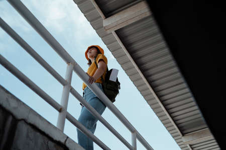 Low angle shot of yellow backpacker woman with green backpack standing at the overpass with roof and...