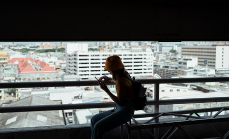 Yellow backpacker woman with green backpack sitting in the parking lot on the high building and using the phone with city view as a background.の写真素材