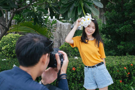 Male photographer take a photo of teenage model girl at the plumeria tree while she hold the flower and camera.の写真素材