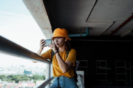 Backpacker girl sitting at the handrail in parking lot of the building taking a photo of the big city.の写真素材