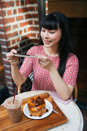 Beautiful asian thai long dark hair woman taking picture of bread cake with smartphone at cafe. Camera eat first.の写真素材