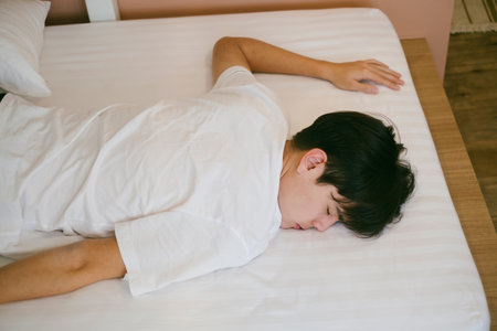 Black hair guy lie down on a stomach on the bed and sleep. Comfy bed in little hotel.の写真素材