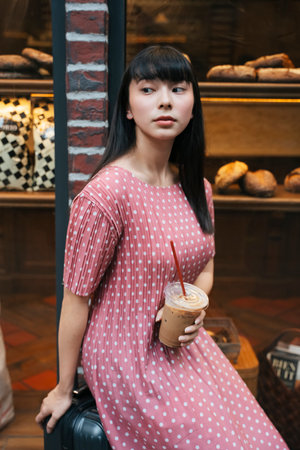 Beautiful asian thai long dark hair woman drinking ice coffee with a straw on street infront of...