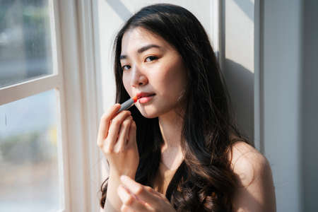 Wavy long hair woman apply red lipstick on her mouth at the window.の写真素材