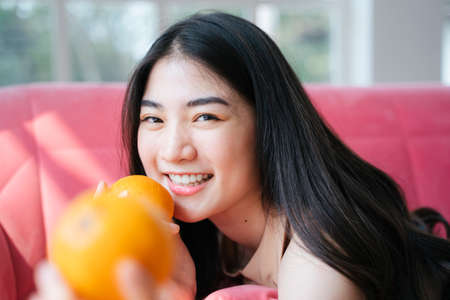 Lively long hair girl lie down on a stomach on the pink sofa, holding the oranges in her hands with bright smile, reach one of orange to the camera.の写真素材