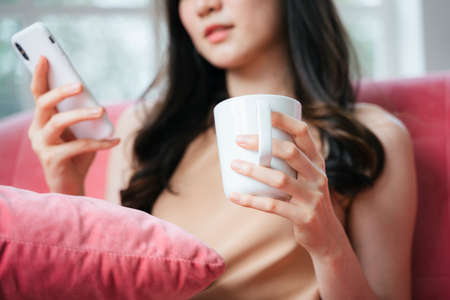 Cropped image of white mug in girl's hand while she sitting on pink sofa and surfing the internet...