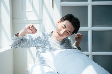 A man sitting on the bed next to the window with morning light, stretching his body to relief the pain.の写真素材