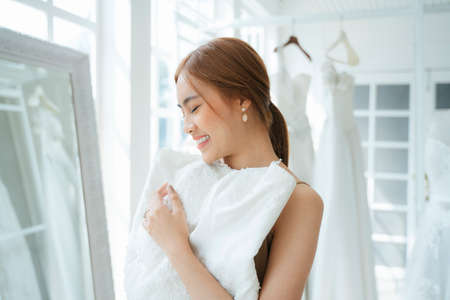 A woman visit wedding studio for wedding dress fitting, she raise the white dress to put over on her body in front of the mirror.の写真素材