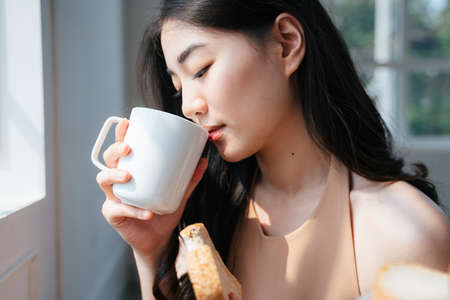 Wavy long hair girl drink hot coffee while sitting next to the window in dining room.の写真素材