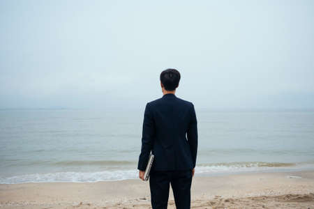 Black hair guy in dark suit holding laptop standing on the beach looking at the sea in gloomy sky.