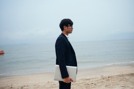 Black hair guy in dark suit holding laptop standing on the beach under unclear sky.の写真素材