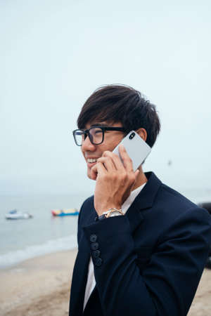 Black hair guy in dark suit standing on the beach and talking on the phone under unclear sky, medium shot.の写真素材