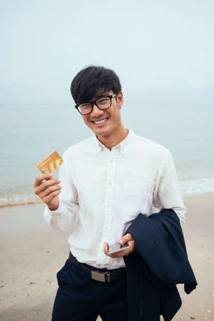Black hair guy in white shirt standing on the beach with gloomy sky and show the credit card, vertical.の写真素材
