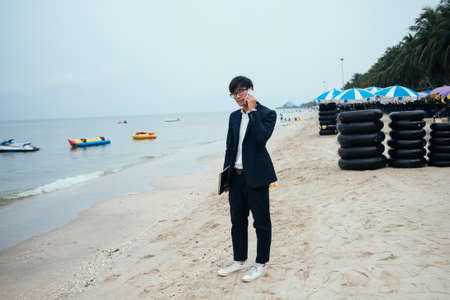 Black hair guy in dark suit standing on the beach and talking with his customer on the phone under unclear sky.の写真素材