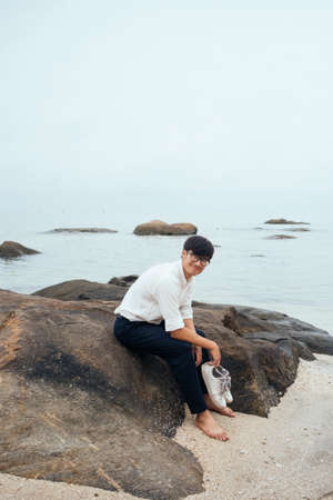 Black hair guy in white shirt take off the shoes and sitting on the rock at the sea, vertical.の写真素材