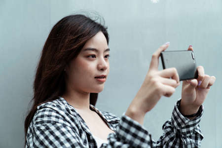 Long hair girl in black plaid standing in front of the wall, raise her smartphone to take a photo.の写真素材