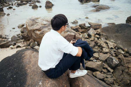 Black hair guy in white shirt sitting on the rock at the sea with bottle in his hand, look at the sea and think about his failing.の写真素材