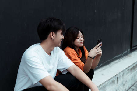 Teenage couple sitting at the black wall, a girl showing her phone to her boyfriend and smile.の写真素材