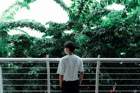 A boy in white t-shirt standing at the handrail of a terrace, trees as a background.