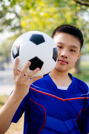 A boy in blue suit give the camera a football while he standing in a park.