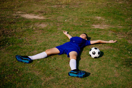 A boy in football suit lie down on the field, spread his arms and legs and sleep with a ball at his...