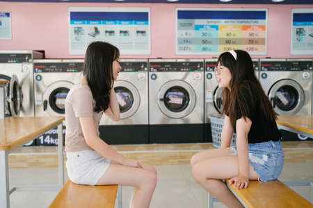 Two girls sitting at the bench in the laundromat, chitcaht and use the smartphone.の写真素材