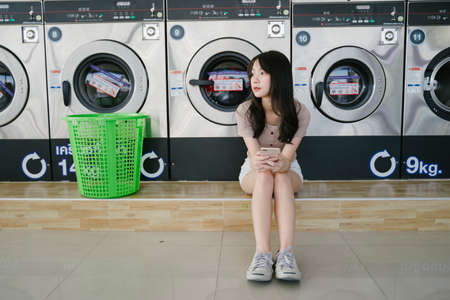 Long hair girl sitting on the floor in front of washing machine waiting for her clothes.の写真素材