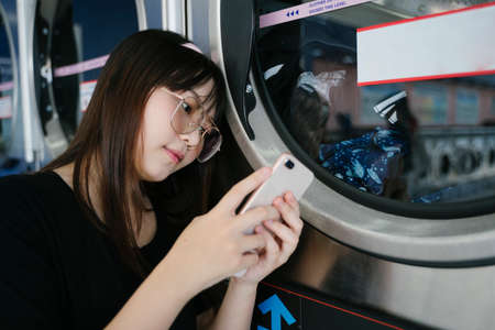Long hair girl wearing glasses use smartphone during waiting for the washing machine, copy space.