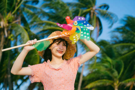 Cute girl in brown hat hold the colorful windmill on the beach in windy day in front of coconut...