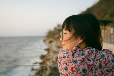 Long hair girl in sunglasses standing at the glasses barricade of the terrace of the building next to the sea and look at the sea in the evening.の写真素材