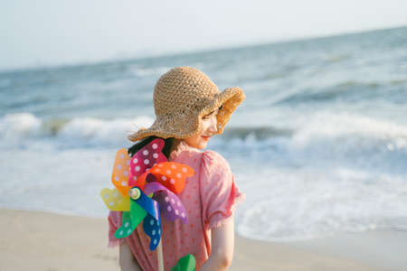 Cute girl standing on the beach and hind the colorful windmill behind herself, backshot.の写真素材