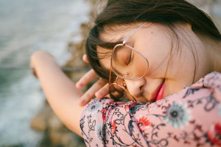 Long hair girl wearing sunglasses close her eyes while prop her head and arms on the glass barricade of the terrace of the weekend house next to the sea.の写真素材