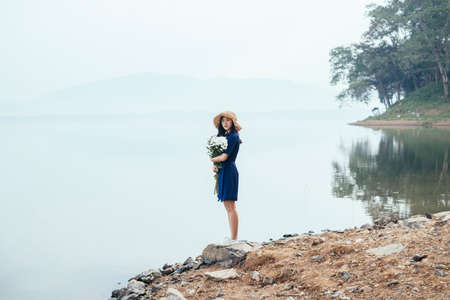 A beautiful Thai girl standing on the rock near by the lake holding the white flowers in her arms looking to the camera.の写真素材