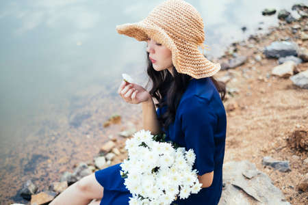 Curly long hair girl enjoy the white flower in her hands while sitting on the rock at the lake.の写真素材