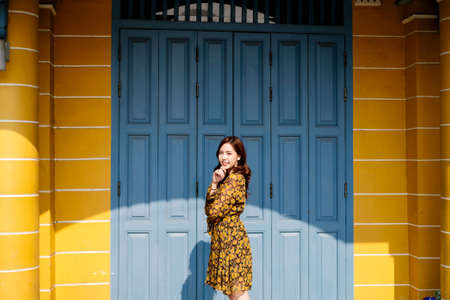 Cute curly long hair girl in yellow dress standing in front of blue folding door and yellow wall, colorful building.の写真素材