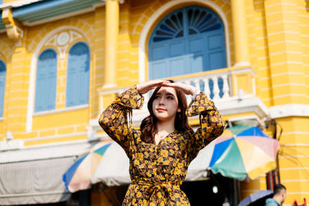 Cute curly long hair girl in yellow dress standing in front of blue and yellow building raise her hands protect the eyes from sunlight to look far away.の写真素材
