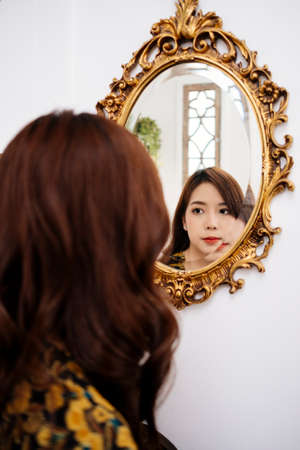 Cute curly long hair girl in yellow dress look at herself in the golden mirror.