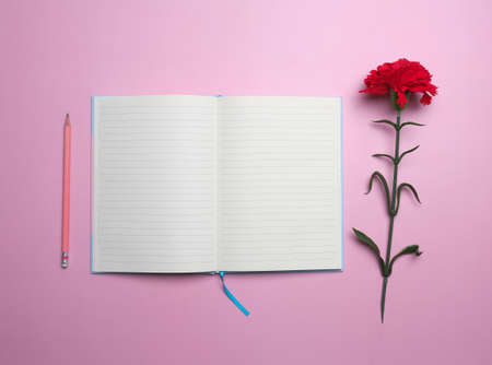 Above view with empty note book, red flower and pencil on pink background.