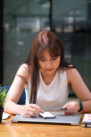 Front view with Asian female in cozy outfit using phone and other hand holding credit card.の写真素材