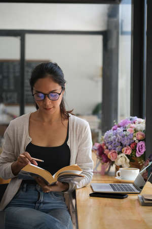 Portrait with Asian woman reading a book while sitting at coffee bar with her laptop and espresso...
