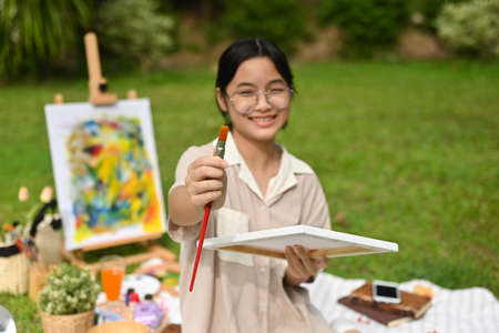 Asian artist girl show the paint brush and focus on brush while sitting on the grass during a picnic in a park, Out door activity concept.の写真素材