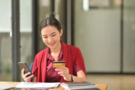 A  woman who wear red shirt holding a credit card and smartphone and sitting on the table have papers, books and devices decorated.の写真素材