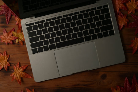 Top view image of a modern laptop computer putting on a wooden table surrounded by dry maple leaves.
