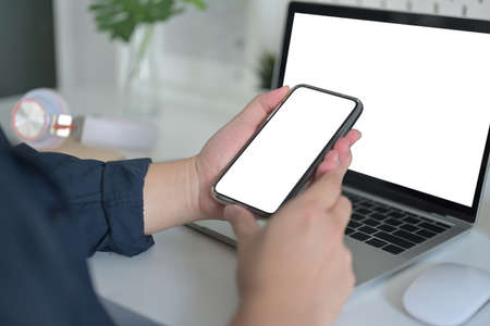 Cropped image of the hand using a white blank screen smartphone in front of laptop.