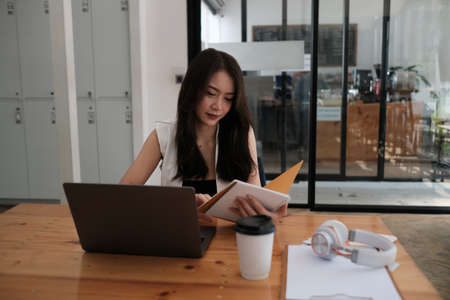 Photo of a beautiful woman taking notes while sitting in front of laptop at the wooden working desk.の写真素材