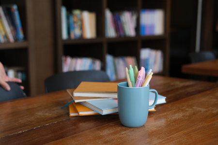 Photo of a cup of pens and stack of books with a bookshelf in the university library as a...